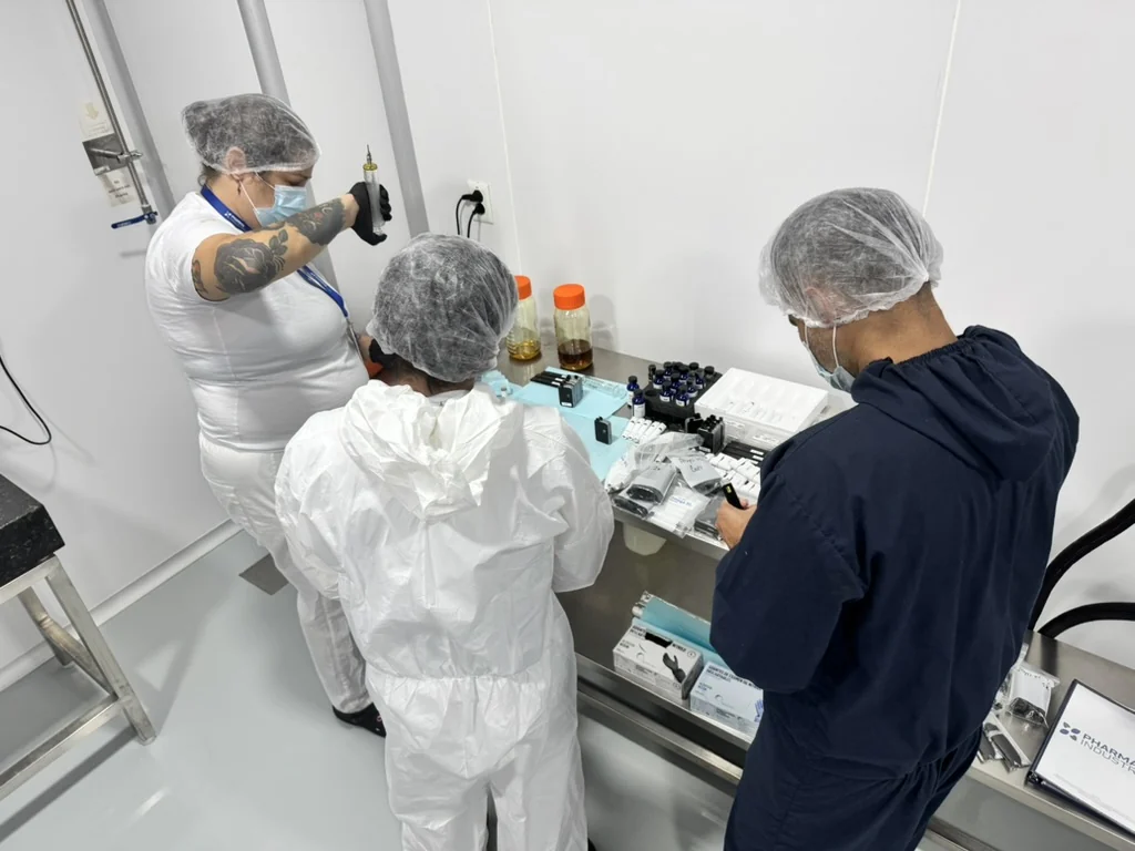 Three gowned operators filling and assembling cartridges at a stainless steel bench in a cleanroom